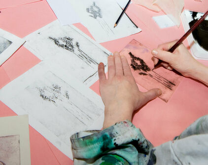 Händer som ristar en bild med torrnål. Runt om ligger papper där den ristade bilden har tryckts.