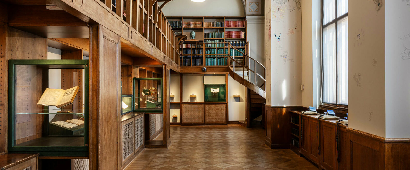 Interiör från ett gammalt bibliotek med böcker i träbokhyllor samt en konstinstallation i glasmontrar och med små böcker på kuddar i bibliotekets fönster.