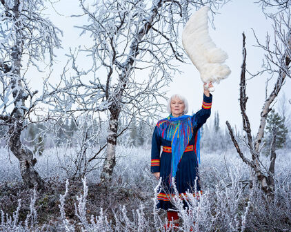 Fotografiskt porträtt föreställande en kvinna i samisk dräkt. Hon står bland björkar i ett snöigt landskap och håller upp en stor fågelvinge.
