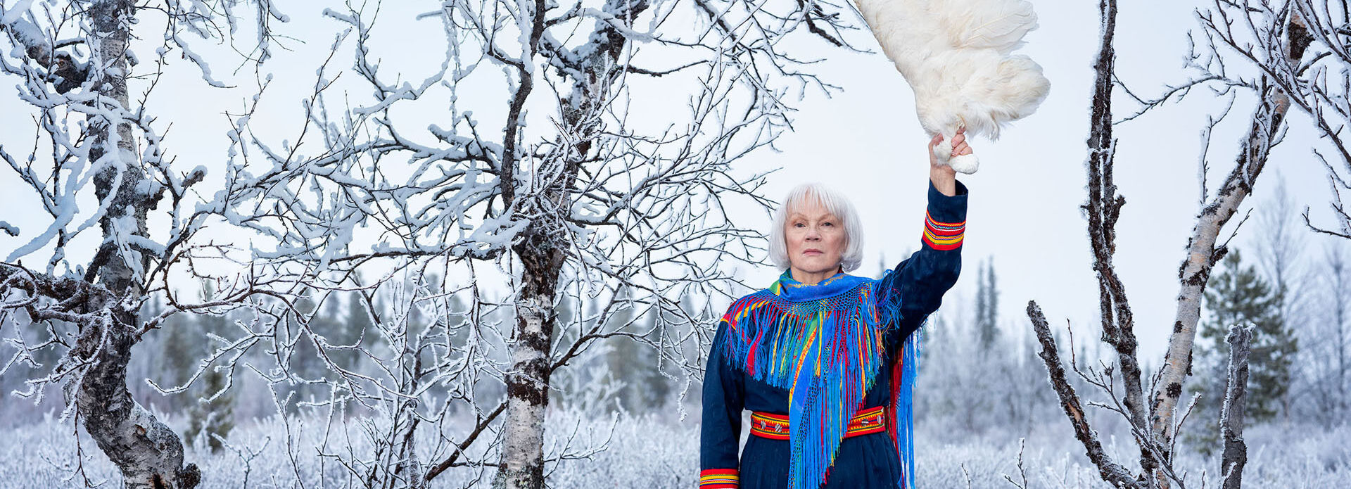 Fotografiskt porträtt föreställande en kvinna i samisk dräkt. Hon står bland björkar i ett snöigt landskap och håller upp en stor fågelvinge.