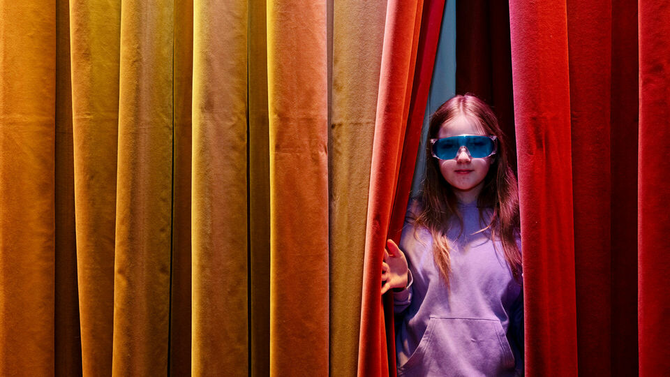 En flicka i knallblå glasögon tittar fram i en glipa i ett färgglatt draperi.