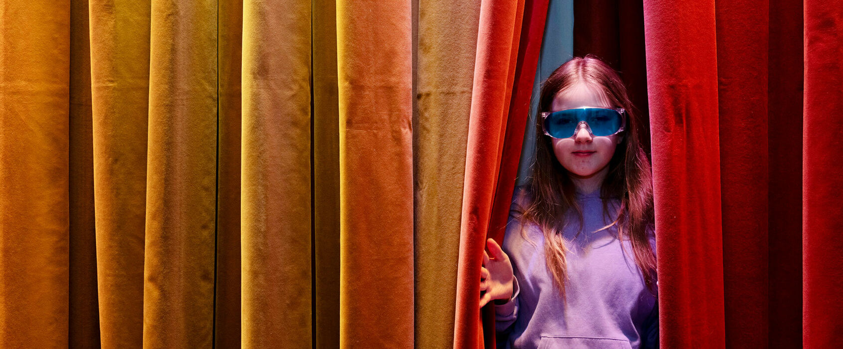 En flicka i knallblå glasögon tittar fram i en glipa i ett färgglatt draperi.