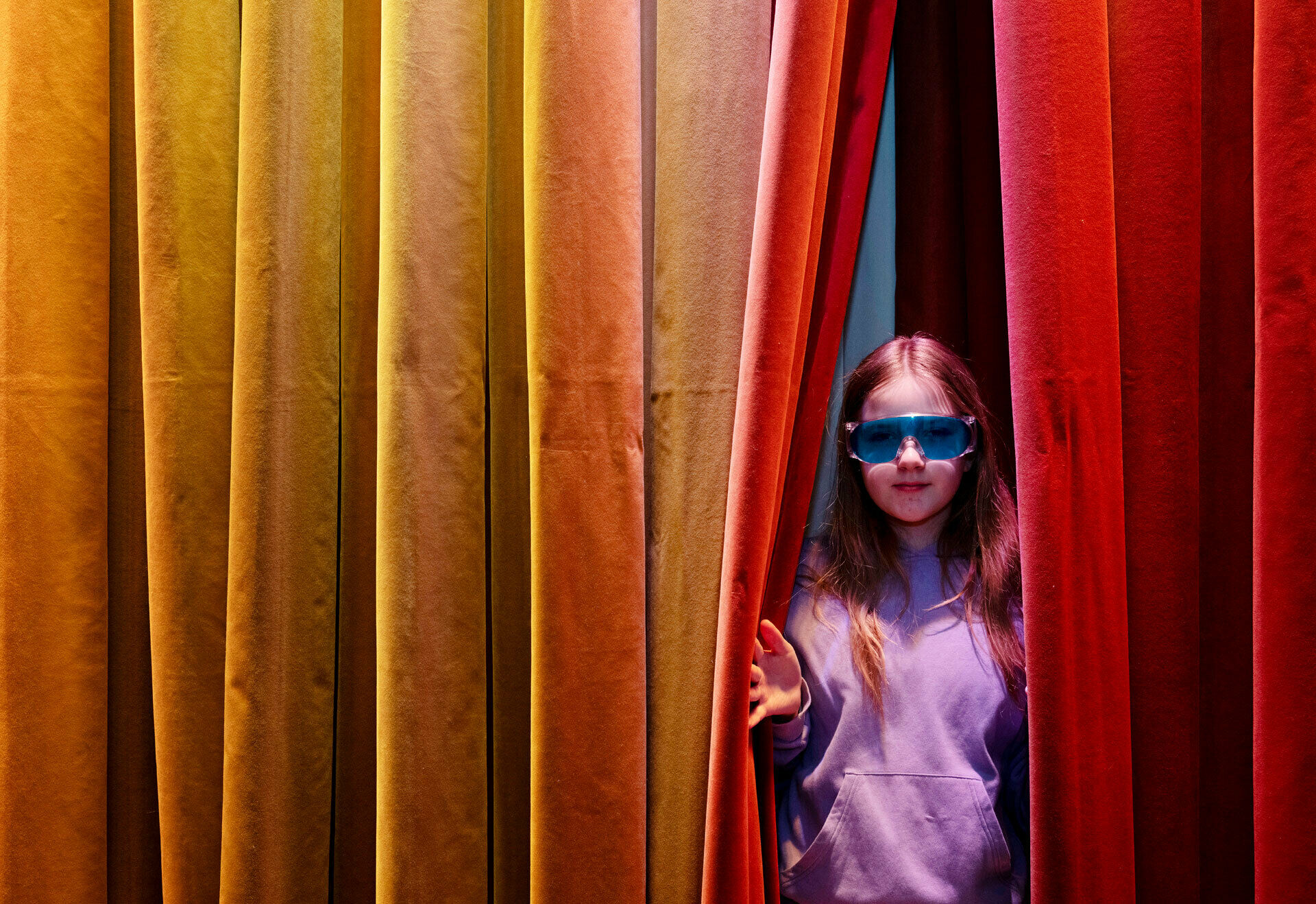 En flicka i knallblå glasögon tittar fram i en glipa i ett färgglatt draperi.