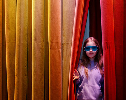 En flicka i knallblå glasögon tittar fram i en glipa i ett färgglatt draperi.