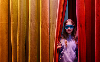 En flicka i knallblå glasögon tittar fram i en glipa i ett färgglatt draperi.