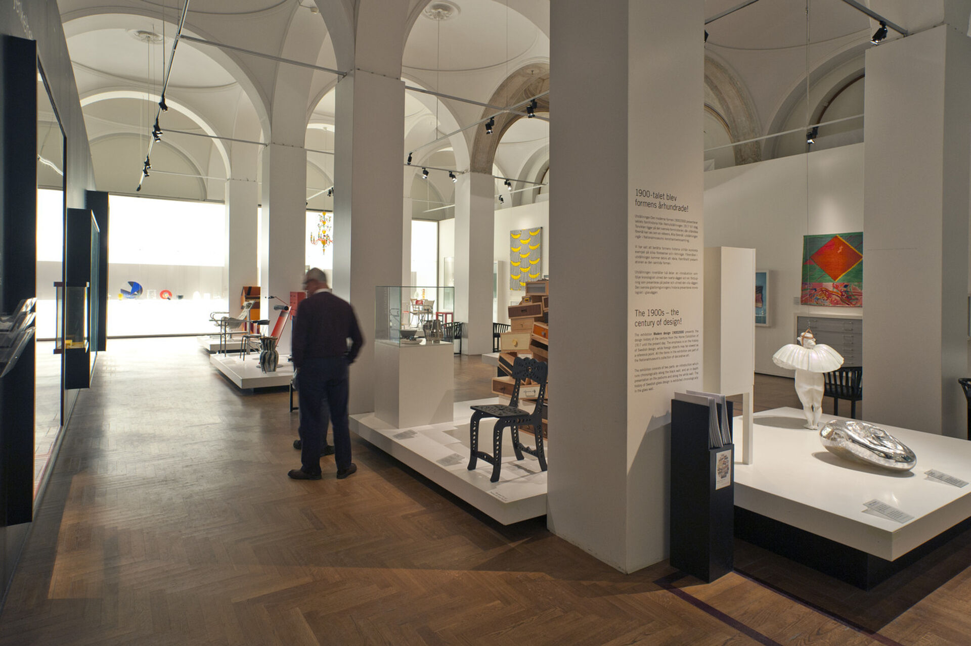 Museisal med vita väggar och brunt parkettgolv. Designföremål på podier vid fyrkantiga höga pelare.