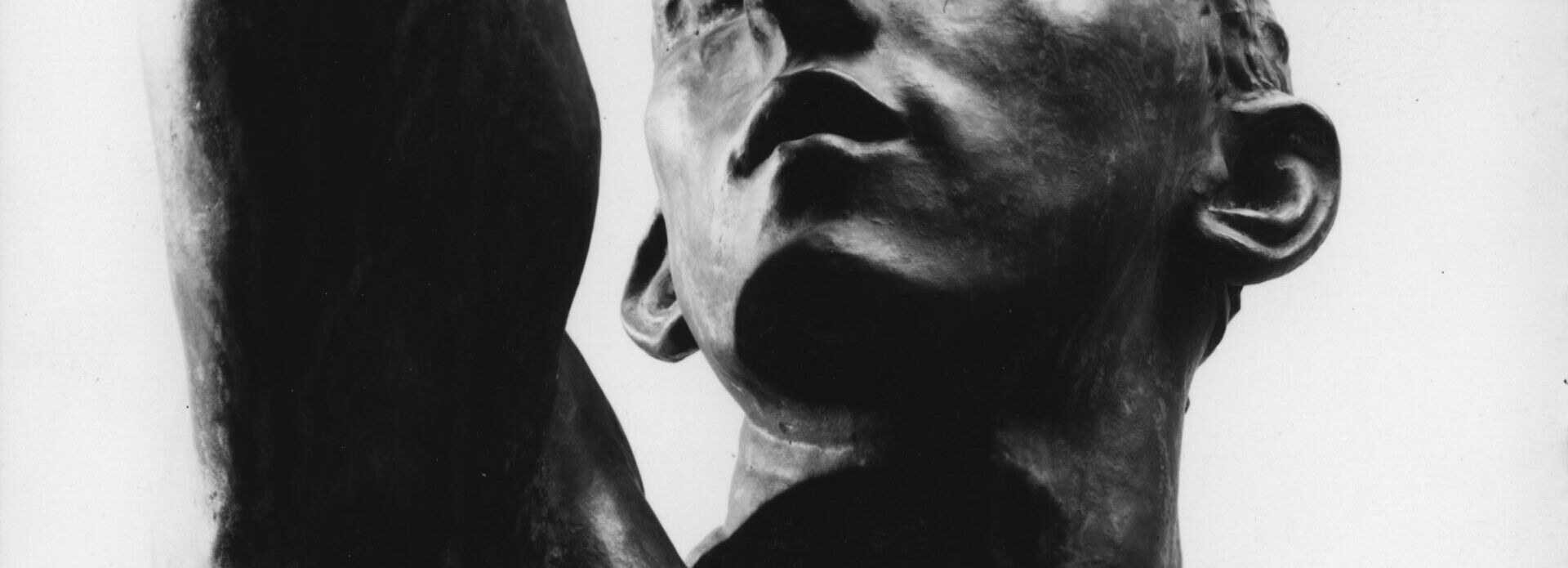 Detalj av bronsskulptur föreställande en med huvudet snett uppåt. Han har ögonen halvt slutna och han håller upp högerarmen och har handen bakom huvudet.