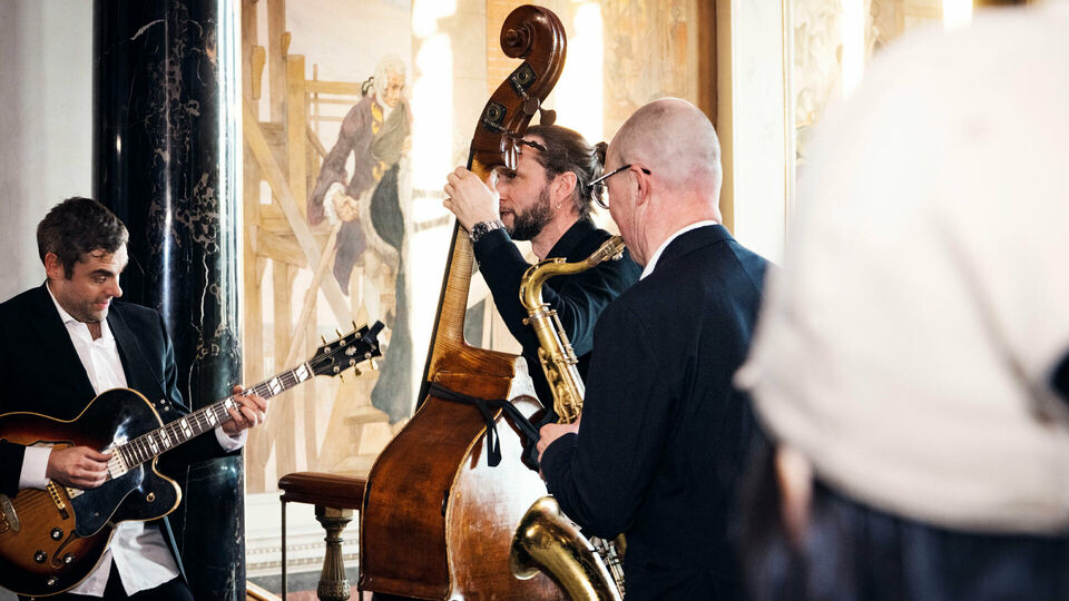 Tre män som står och spelar på gitarr, kontras och saxofon i en storslagen trapphall med målade fresker.