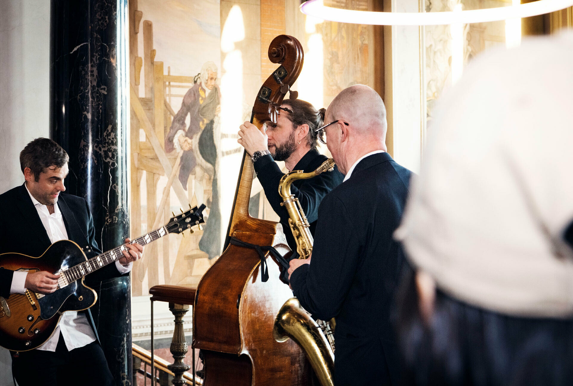 Tre män som står och spelar på gitarr, kontras och saxofon i en storslagen trapphall med målade fresker.