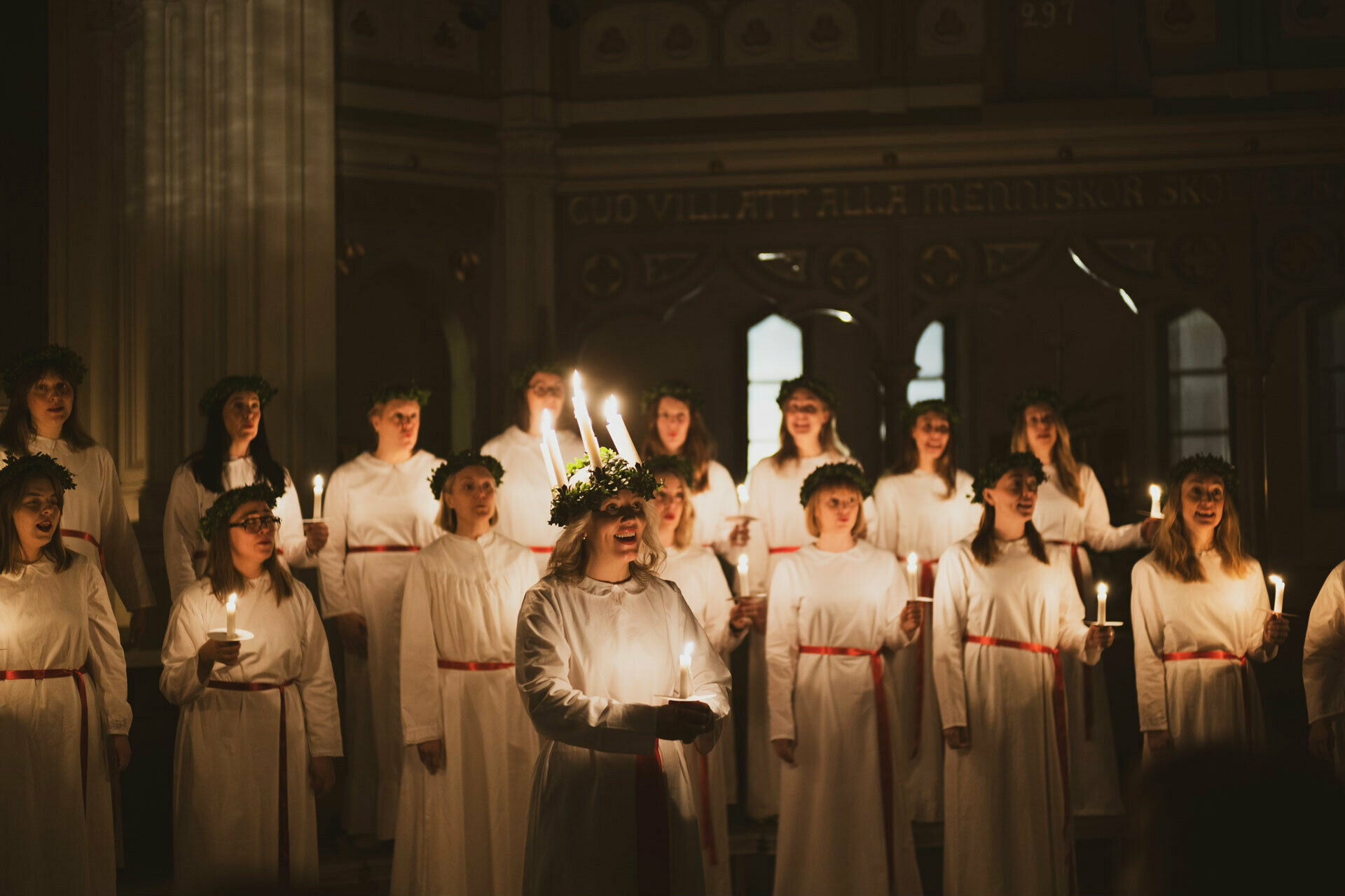 Kvinnor i luciatåg i miljö som ser ut att vara en kyrka.
