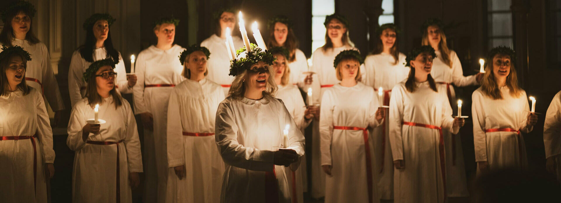 Kvinnor i luciatåg i miljö som ser ut att vara en kyrka.