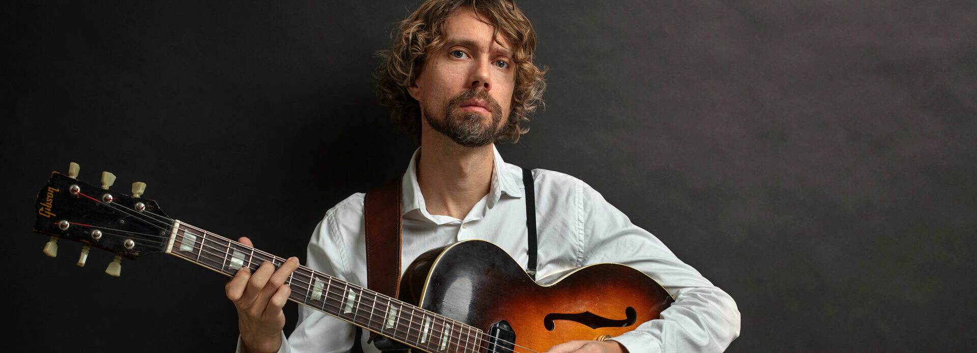 Fotografi av en man i vit skjorta och svarta byxor som håller en gitarr i famnen.