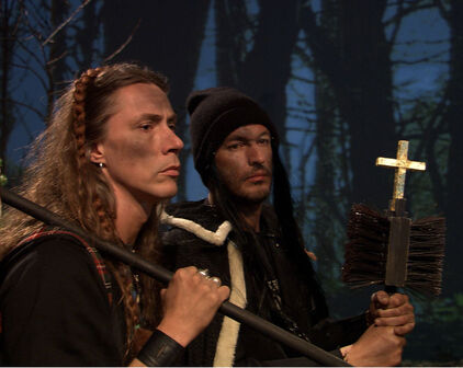 Stillbild från film. Två män i en skog. Den ena håller i ett kors fästad på ett par sopborstar, den andre håller en stång som en slags fana är fästad på.