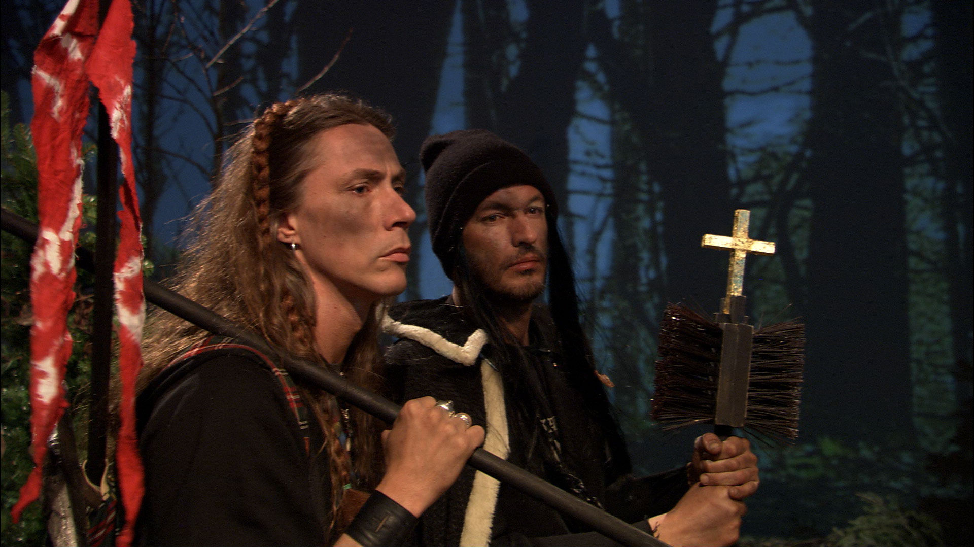Stillbild från film. Två män i en skog. Den ena håller i ett kors fästad på ett par sopborstar, den andre håller en stång som en slags fana är fästad på.