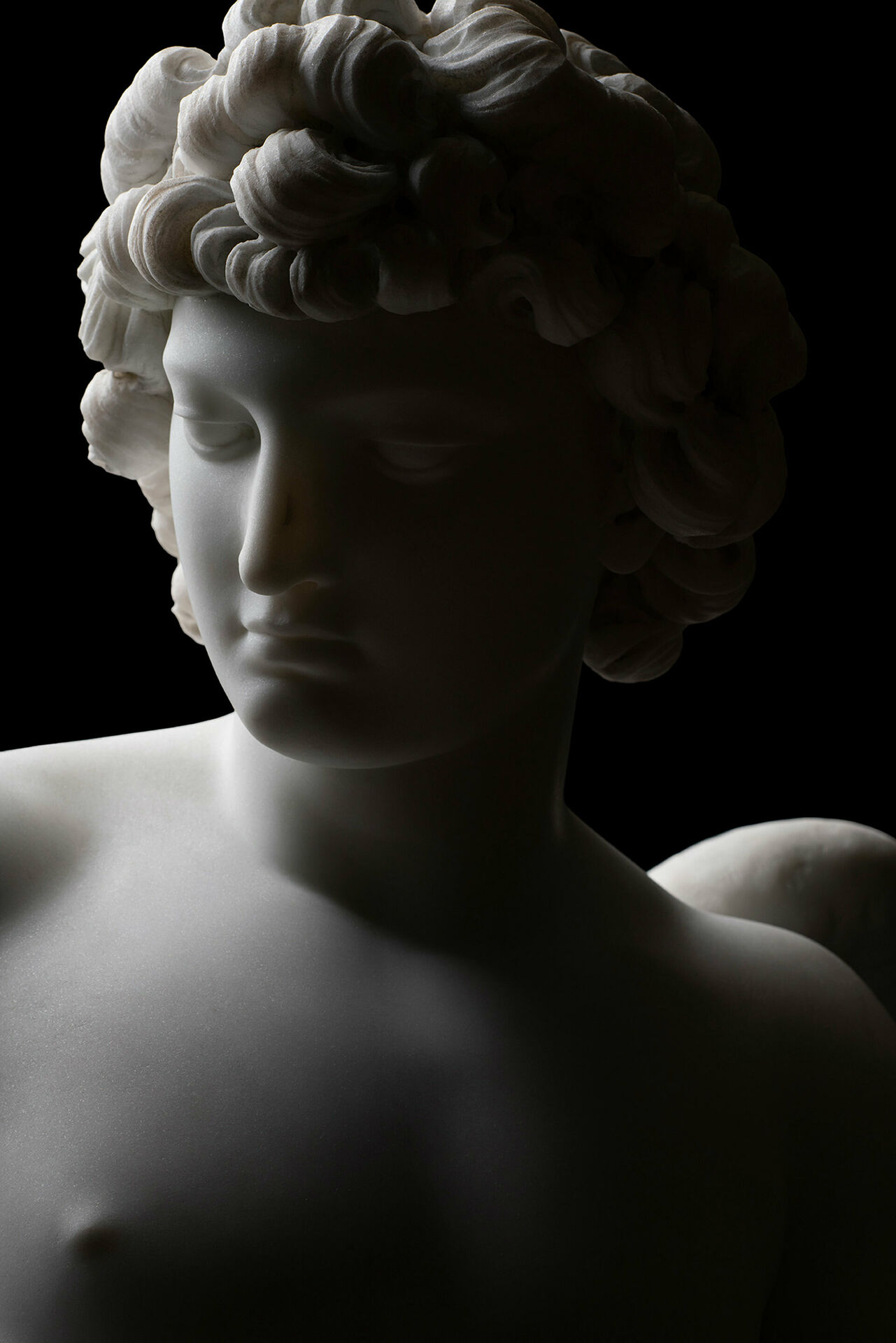 Detalj från marmorskulptur, huvudet på en ung man med lockigt hår, sedd snett från sidan.