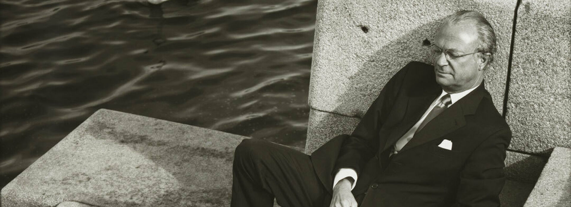 En man, Sveriges kung Carl XVI Gustav, som sitter i en trappa ner mot vattendrag.