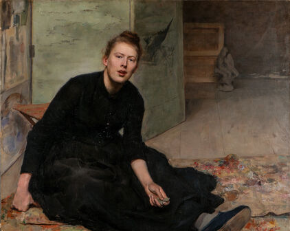 Porträtt från ca 1900 av en kvinna i svart klänning som sitter på golvet. I handen och runt omkring sig har hon penslar och annat konstnärsmateriel.