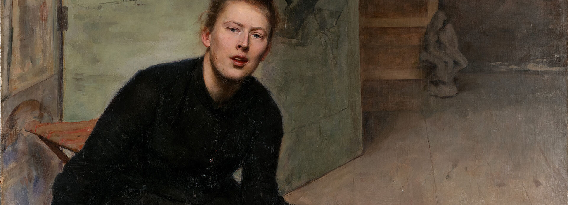 Porträtt från ca 1900 av en kvinna i svart klänning som sitter på golvet. I handen och runt omkring sig har hon penslar och annat konstnärsmateriel.