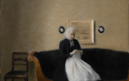 Äldre kvinna sittande i soffa med ett handarbete i händerna.