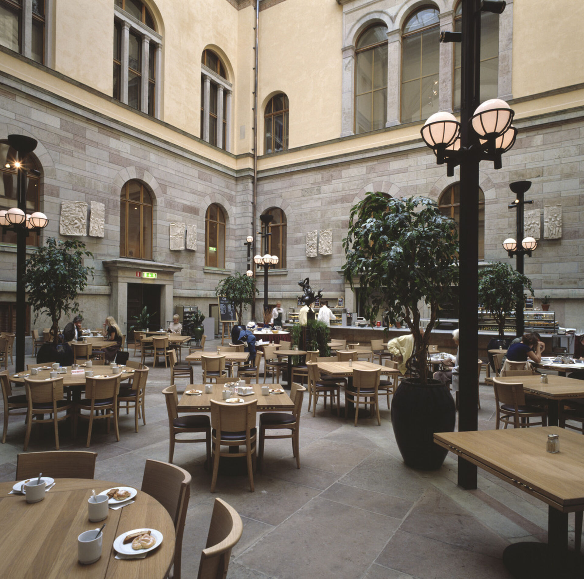 Stort inomhustorg med stora gröna växter. Grått stengolv, gulaktiga väggar, belysning på höga stolpar. Stolar och bord.