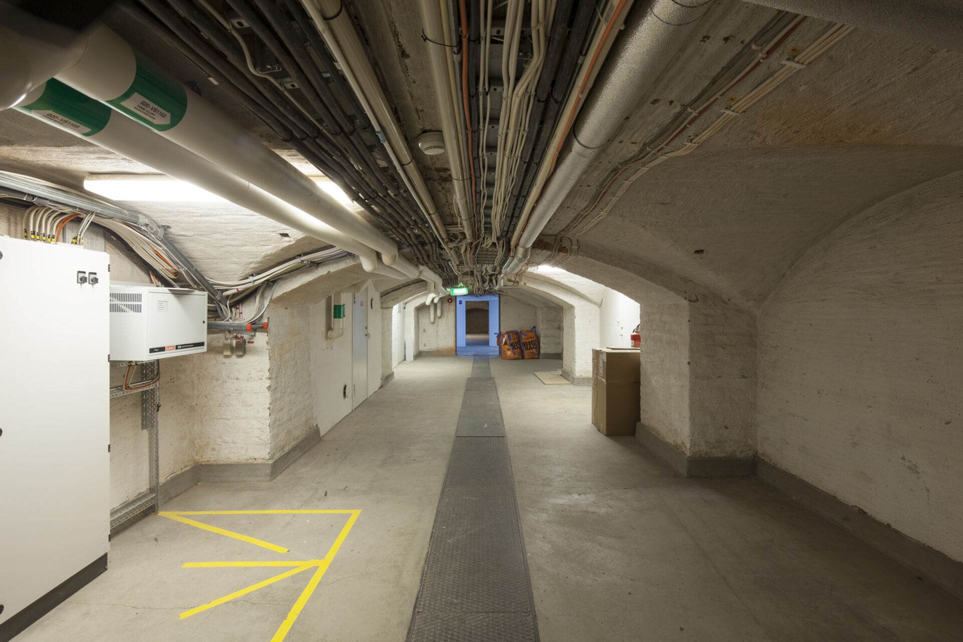 Källargång med välvt tak. Rör och kablar i taket. Maskiner vid väggen.