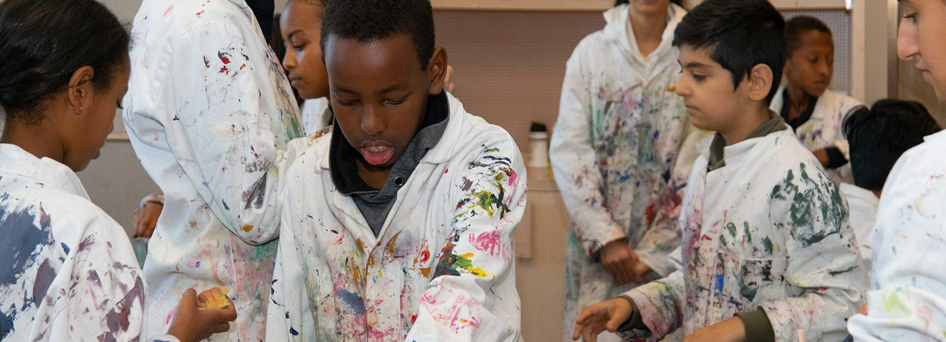 Barn i målarrockar som botaniserar bland penslar som står i en burk i en ateljé.