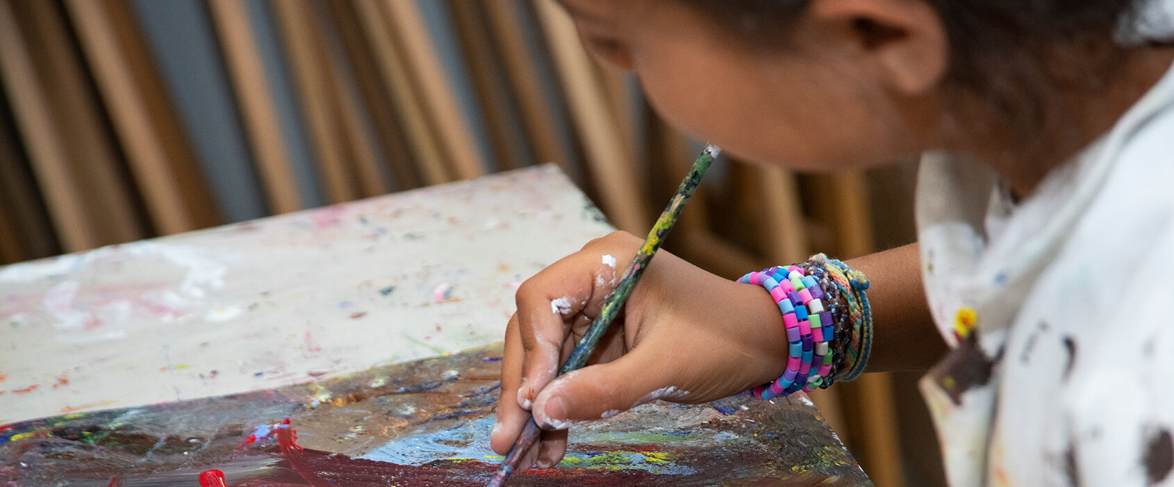 Ett barn som sitter vid ett bord i en ateljé och målar med akrylfärg i starka kulörer.