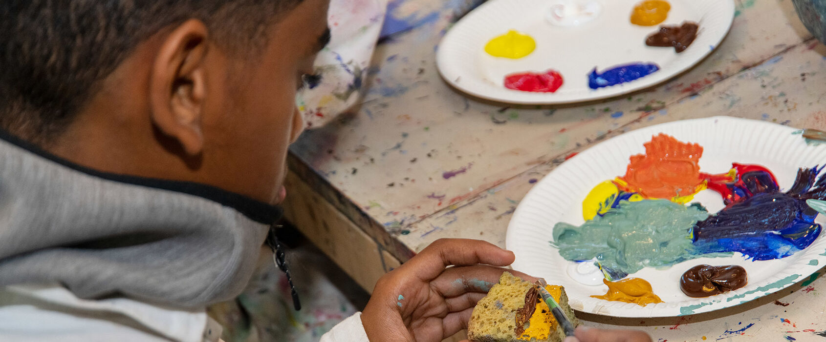 Ett barn som sitter vid ett bord i en ateljé och duttar akrylfärg på en svamp.