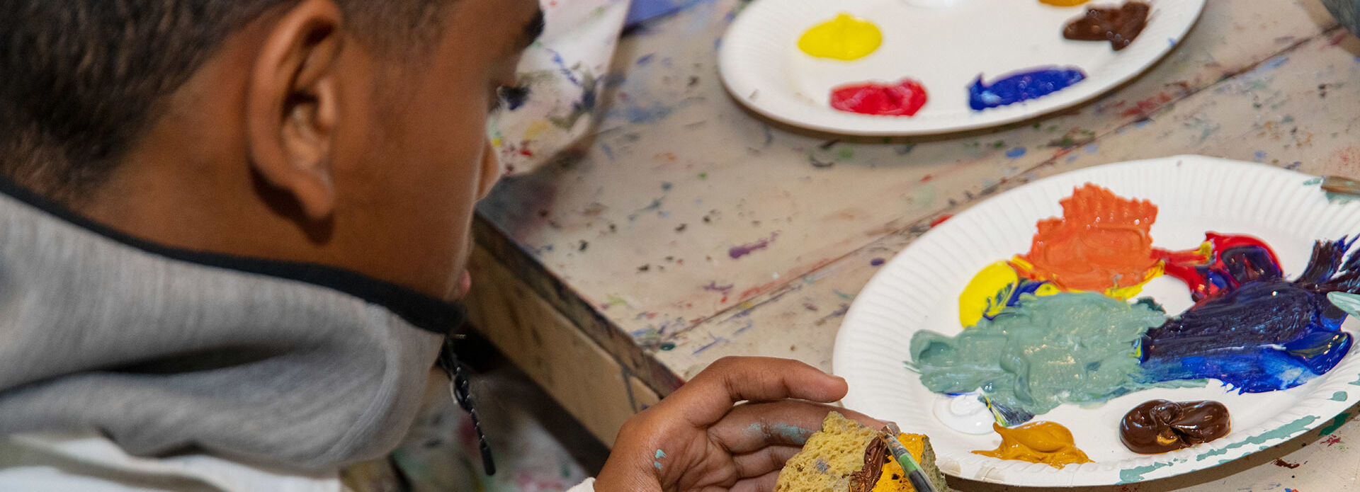 Ett barn som sitter vid ett bord i en ateljé och duttar akrylfärg på en svamp.