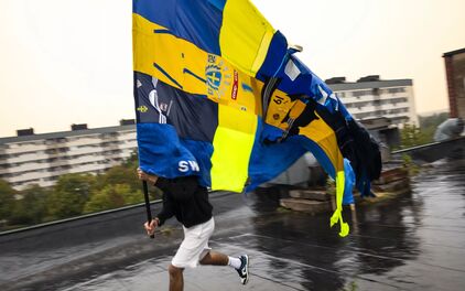 En person springer på ett hustak med en stor blågul flagga på en pinne i handen. Flaggan vajjar i vinden och på den sitter diverse sportbudskap.
