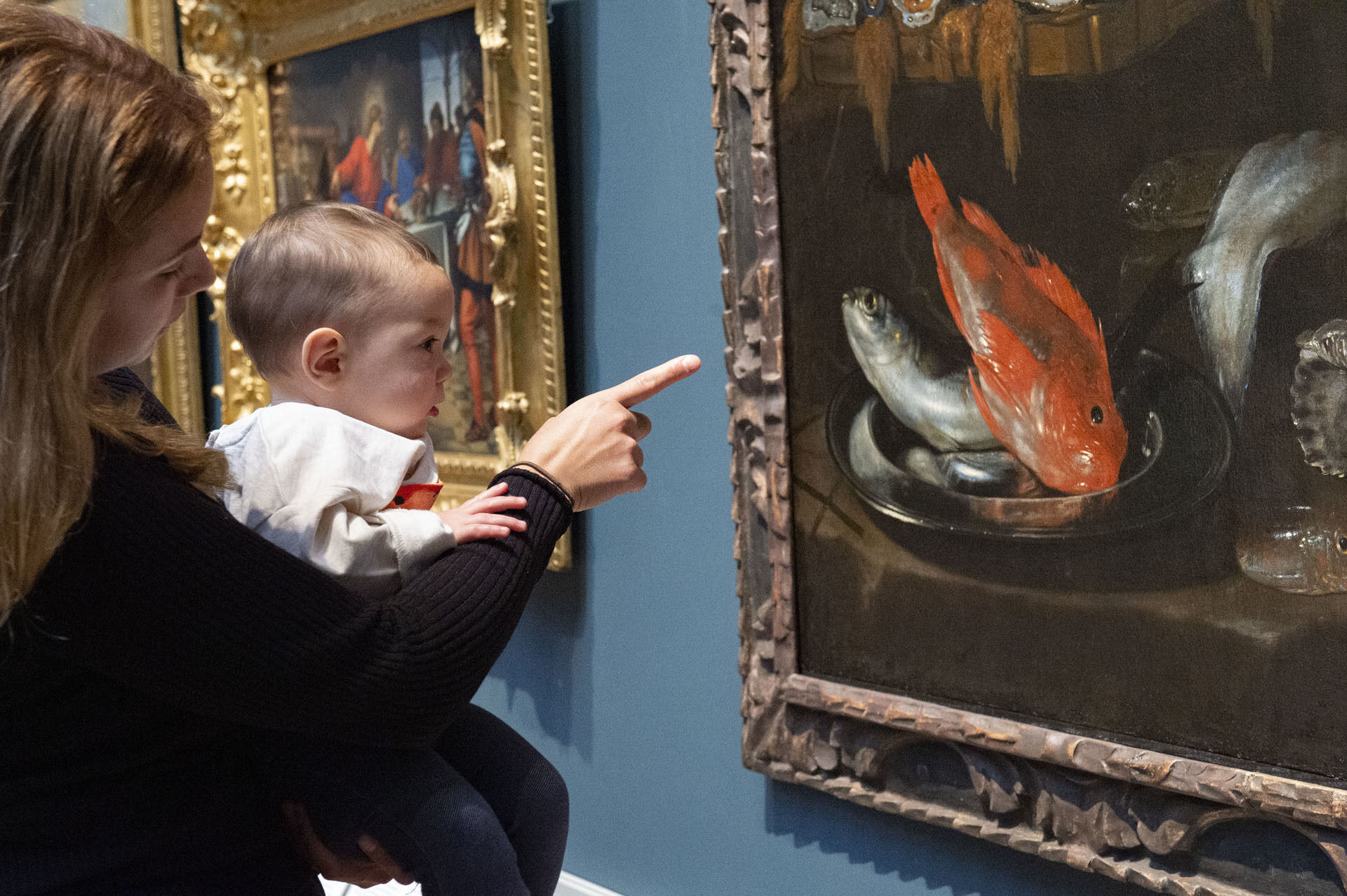 En kvinna håller en baby i famnen och tillsammans tittar de på en tavla med en röd fisk.