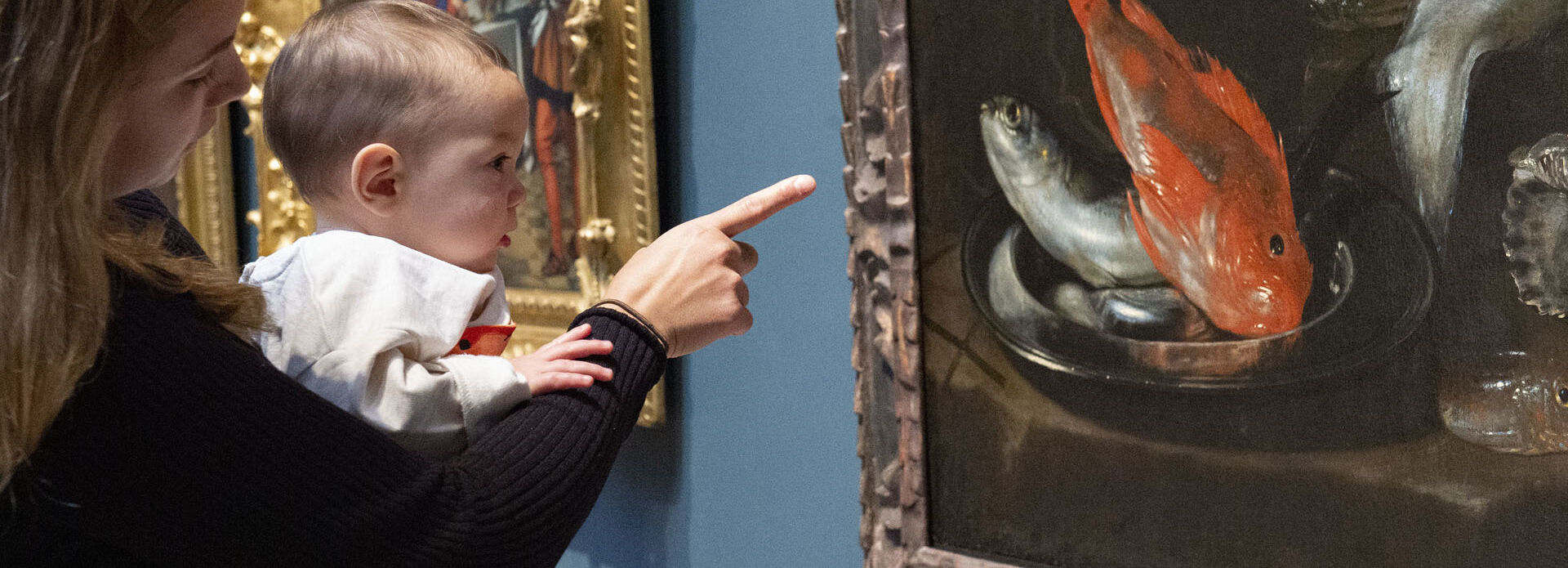 En kvinna håller en baby i famnen och tillsammans tittar de på en tavla med en röd fisk.
