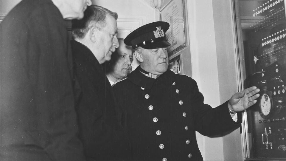 En man i uniform pekar på elektriska reglage och knappar. Tre män tittar på.