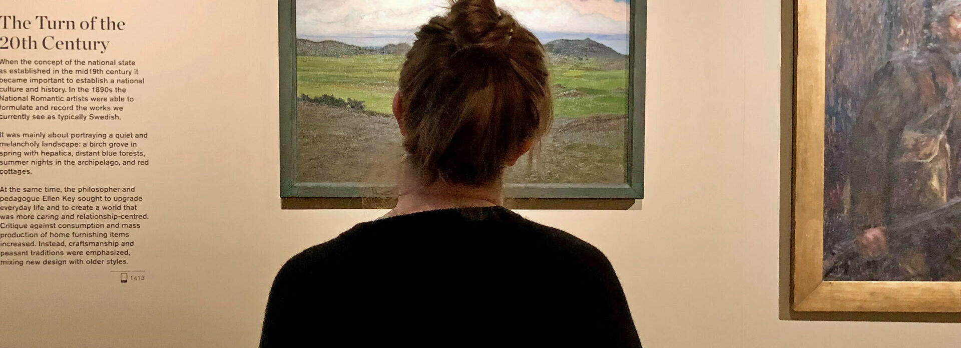 En kvinna som sitter med ryggen åt betraktaren. Hon tittar på en landskapsmålning som hänger på en vägg.