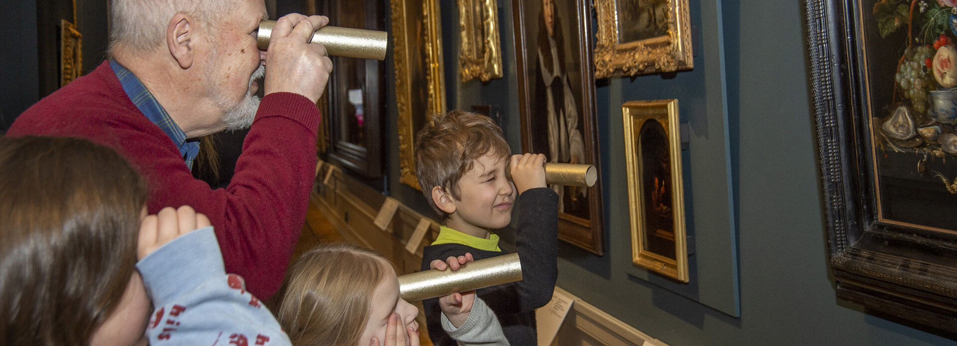 En äldre man och tre barn tittar på tavlor i en museisal med hjälp av hemmagjorda kikare, eller tittinstrument.