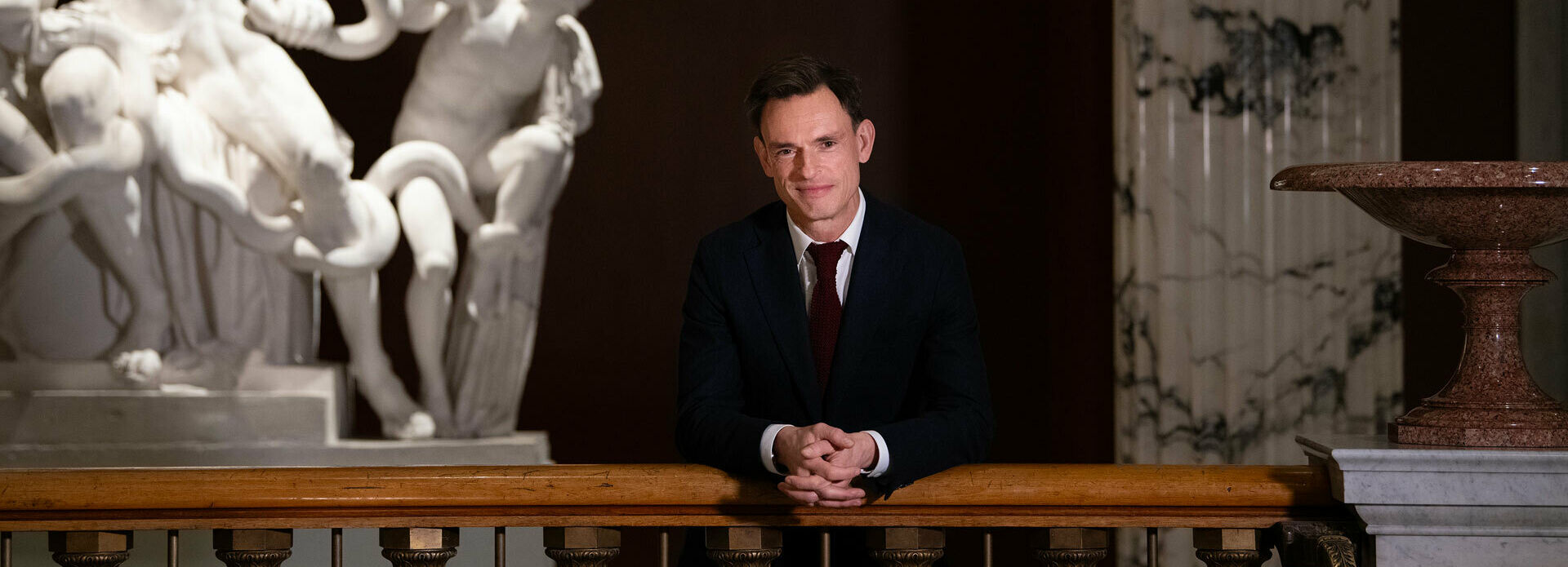 Foto av en man som står lutad mot ett trappräcke i en trapphall. Intill honom en avgjutning av en antik skulptur.