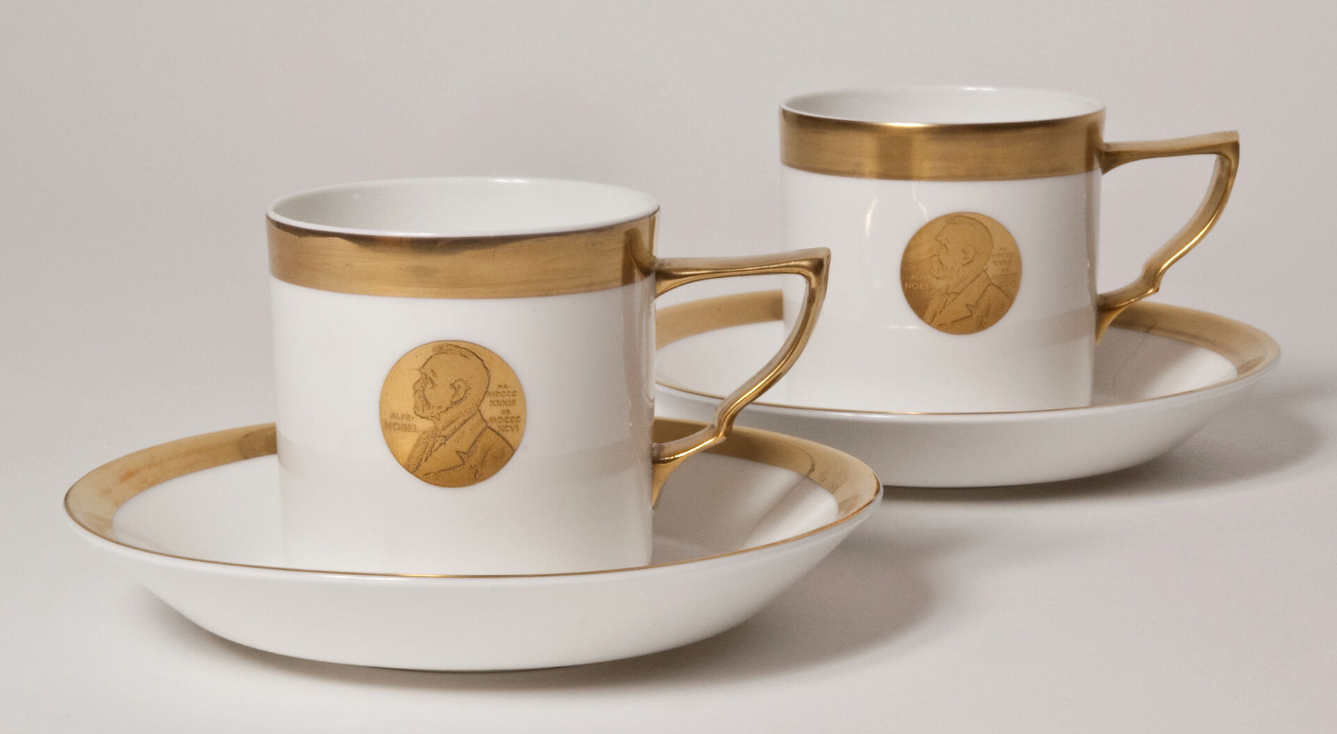 Två kaffekoppar i vitt och guld med en guldmedaljong föreställande Alfred Nobel mitt på koppen.