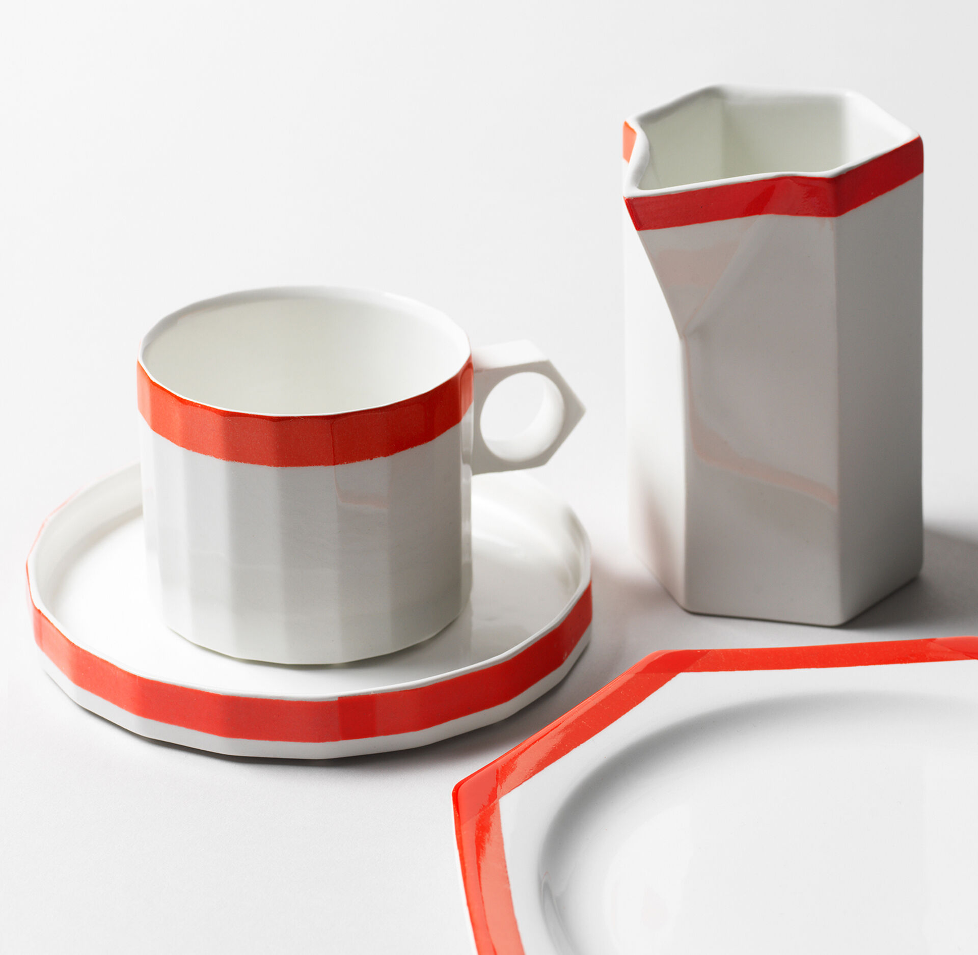 Kaffekopp, tallrik och mjölkkanna i vit glasyr med röd kant.
