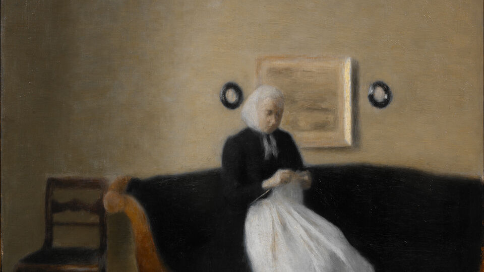 Äldre kvinna sittande i soffa med ett handarbete i händerna.