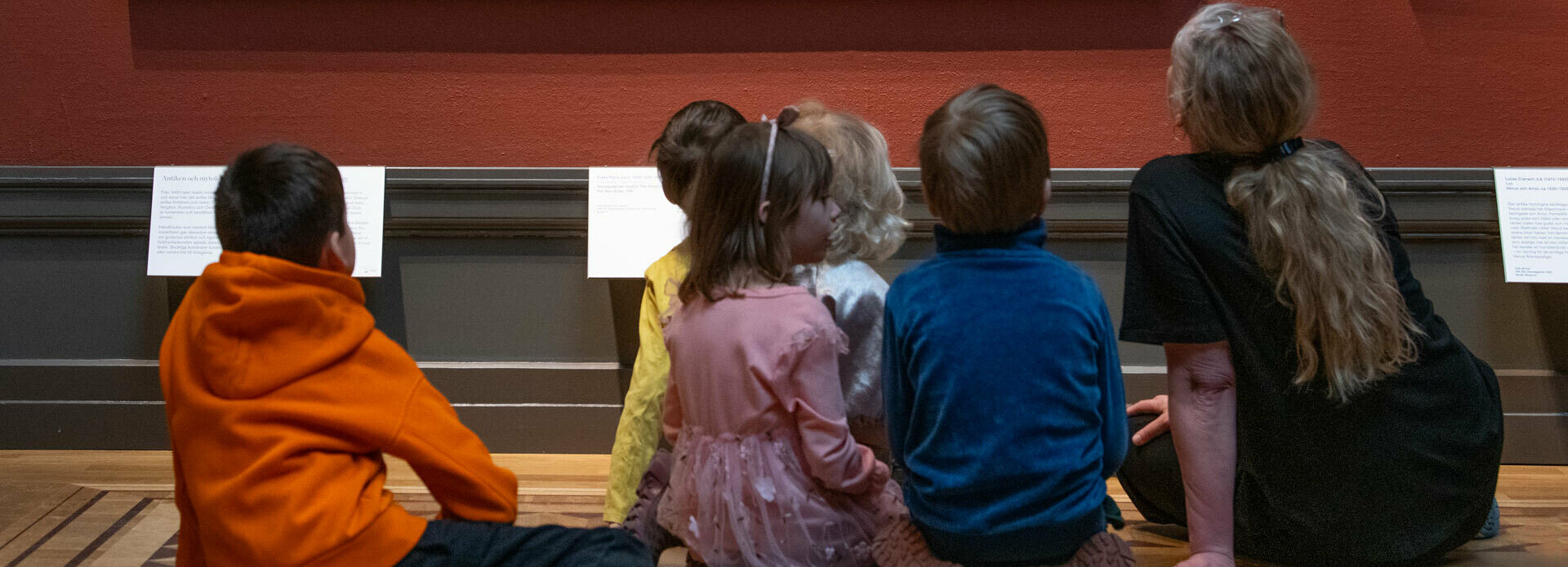 Några barn och en vuxen kvinna sitter på golvet i ett museum och tittar på en målning som hänger på väggen.