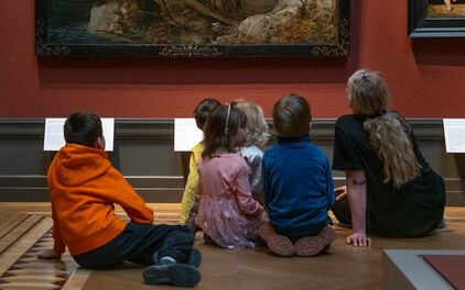 Några barn och en vuxen kvinna sitter på golvet i ett museum och tittar på en målning som hänger på väggen.