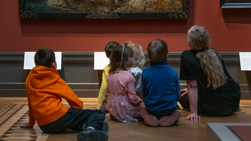 Några barn och en vuxen kvinna sitter på golvet i ett museum och tittar på en målning som hänger på väggen.