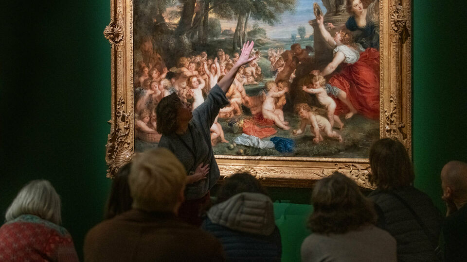 Vuxna personer sitter och tittar på en målning på en museivägg. En kvinna står upp oh pekar på målningen, hon ser ut att berätta om den.