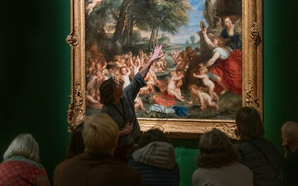 Vuxna personer sitter och tittar på en målning på en museivägg. En kvinna står upp oh pekar på målningen, hon ser ut att berätta om den.