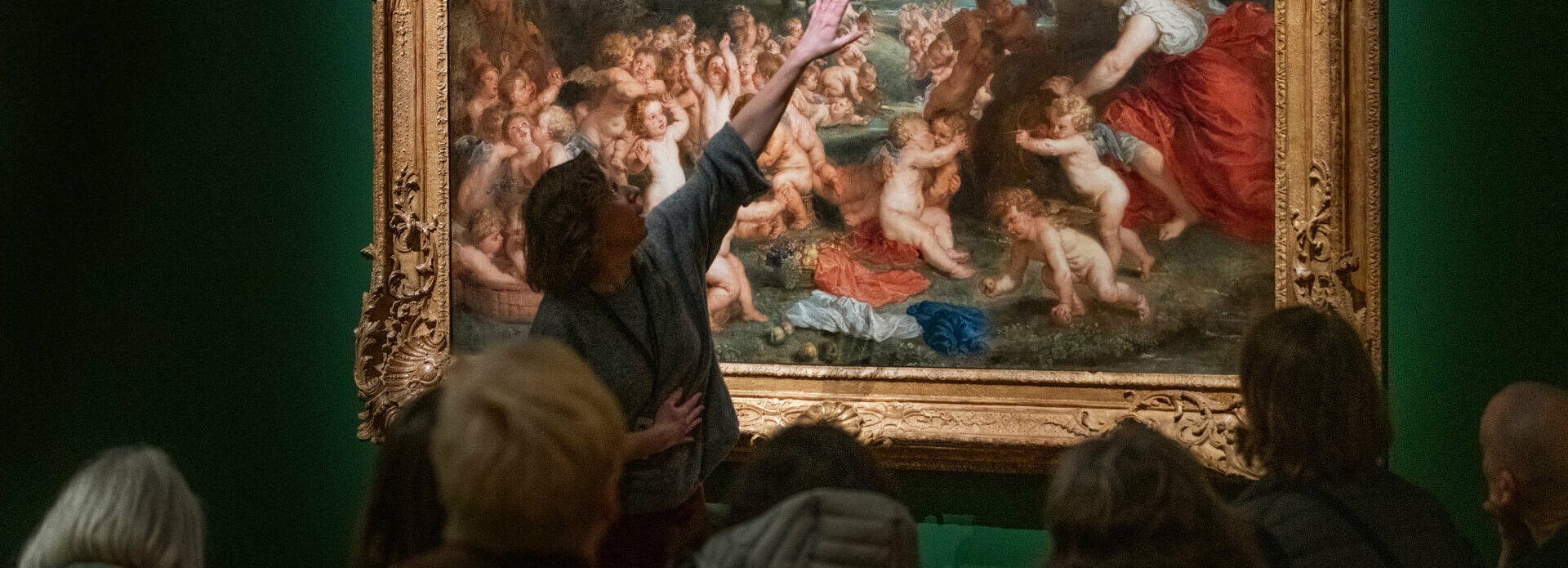 Vuxna personer sitter och tittar på en målning på en museivägg. En kvinna står upp oh pekar på målningen, hon ser ut att berätta om den.