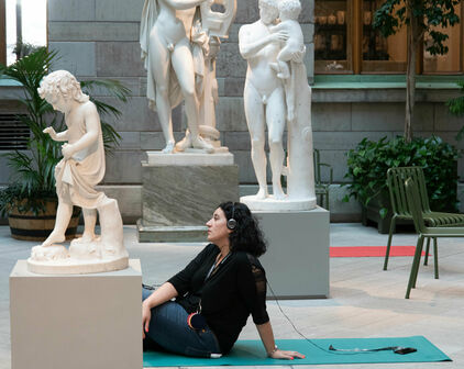 En kvinna sitter på en yogamatta i en skulpturgård. Hon har hörlurar på huvudet och ser ut att vara försjunken i tankar.