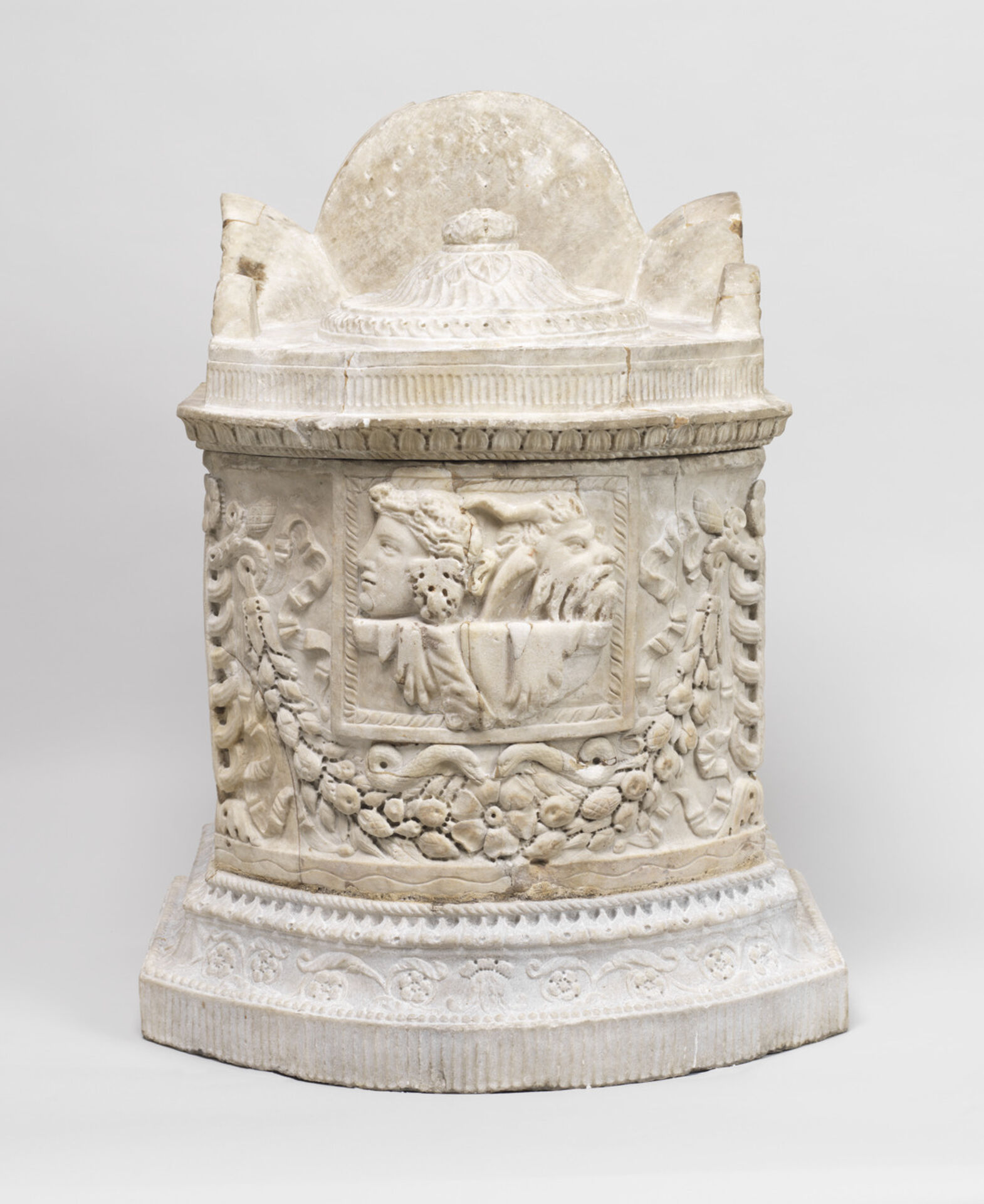 En vit marmorskulptur med rundad överdel och rikt utsmyckad sockel. På framsidan syns två ansikten i relief, omgivna av hängande blomster- och fruktgirlander. Skulpturen har dekorativa bårder och en bred, ornamenterad bas.