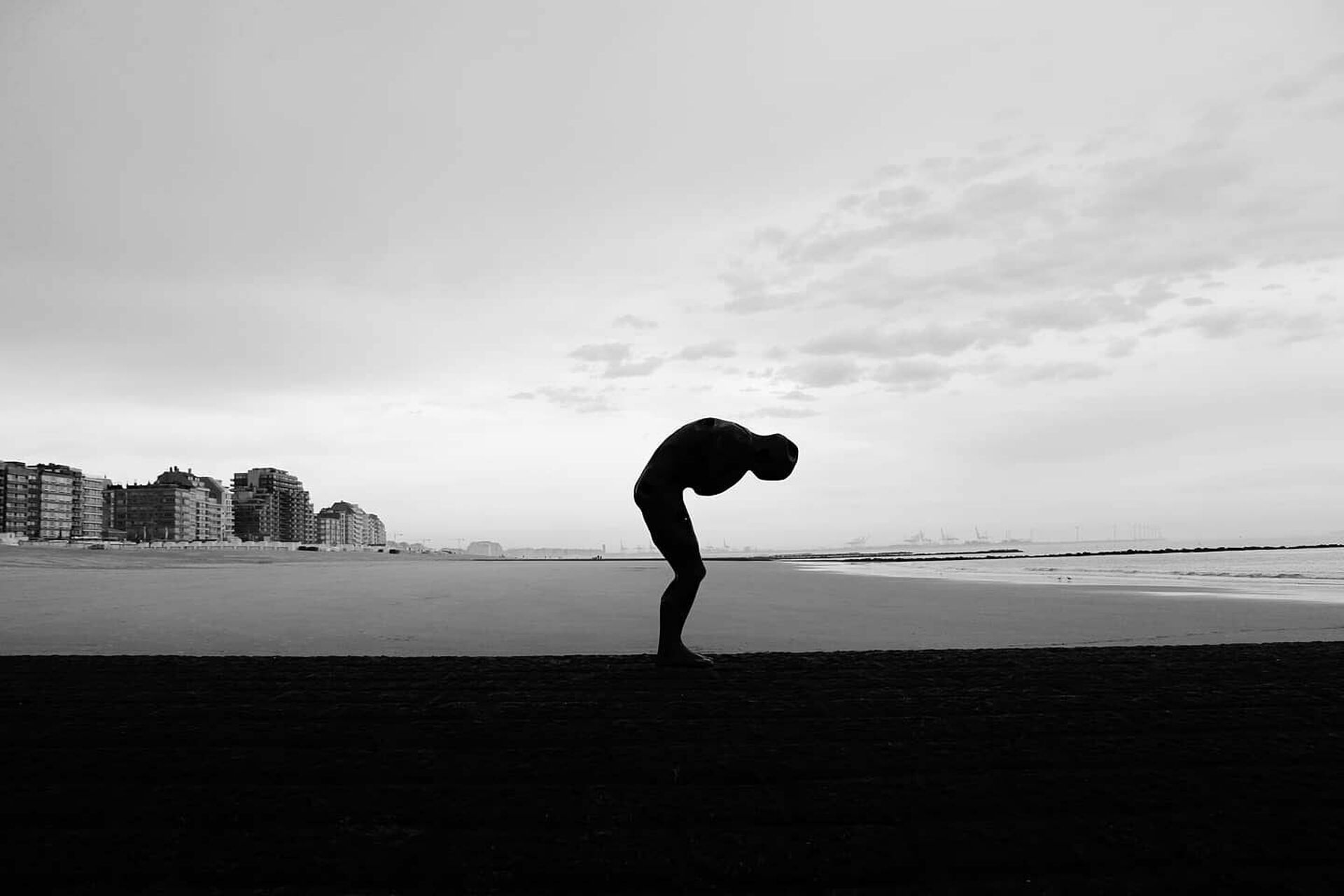 Poetisk bild av en människa som står hopkurad på en öde strand. I fonden syns höga hus och ute i havet skymtar kranar för varvsverksamhet.