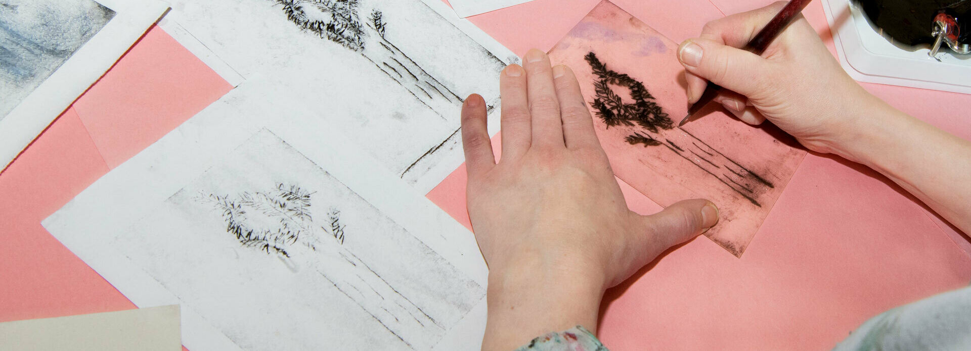 Händer som ristar en bild med torrnål. Runt om ligger papper där den ristade bilden har tryckts.