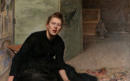 Porträtt från ca 1900 av en kvinna i svart klänning som sitter på golvet. I handen och runt omkring sig har hon penslar och annat konstnärsmateriel.